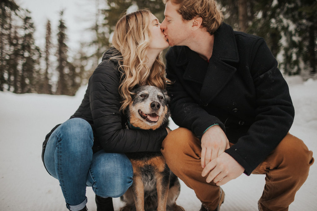 couple kissing in winter wonderland while cuddling their Australian Shepard dog