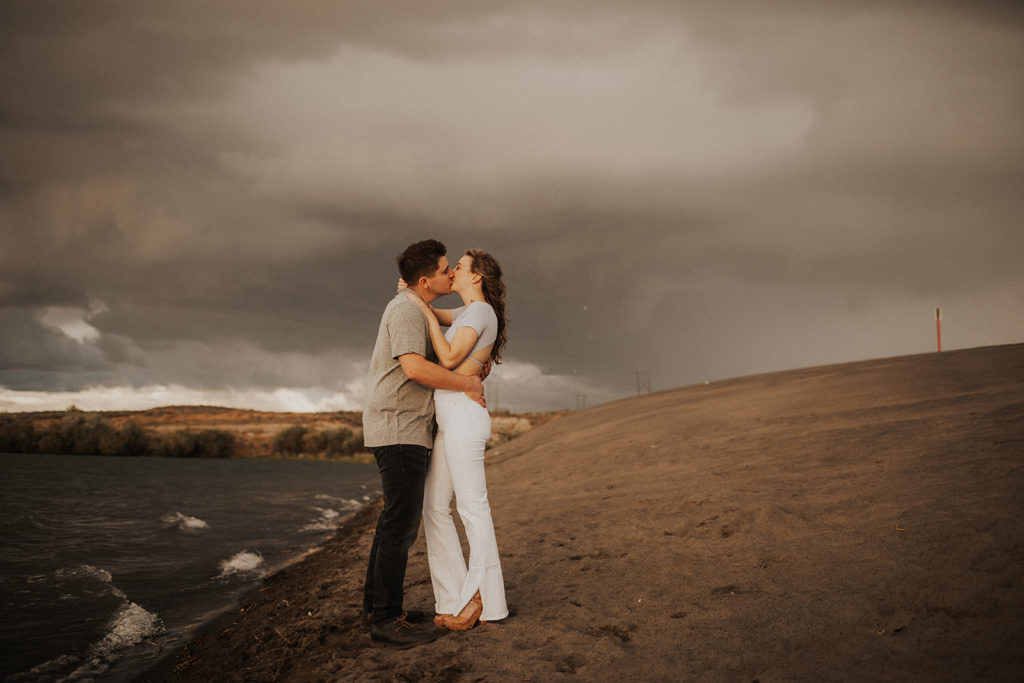 engagement photos at sand dunes - couple kissing on the beach, her in white pants and light blue crop top and him in black pants and grey shirt