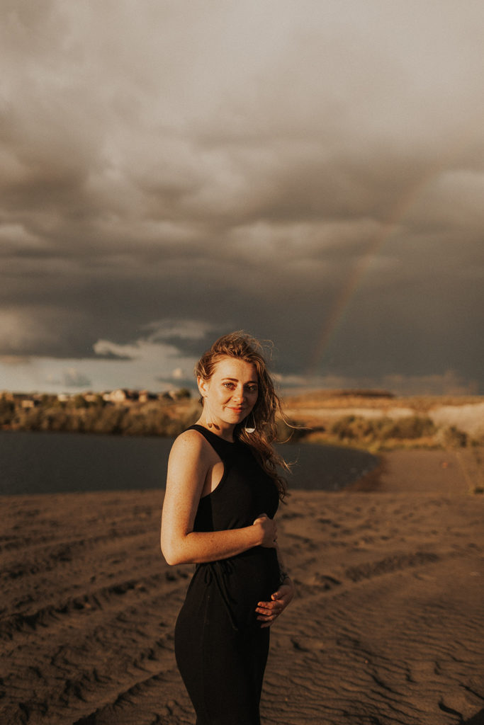 woman in black dress caresses baby bump at sand dunes with rainbow in the background