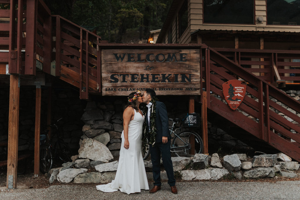 bride and groom kiss in front of stehekin (small town in national forest) sign