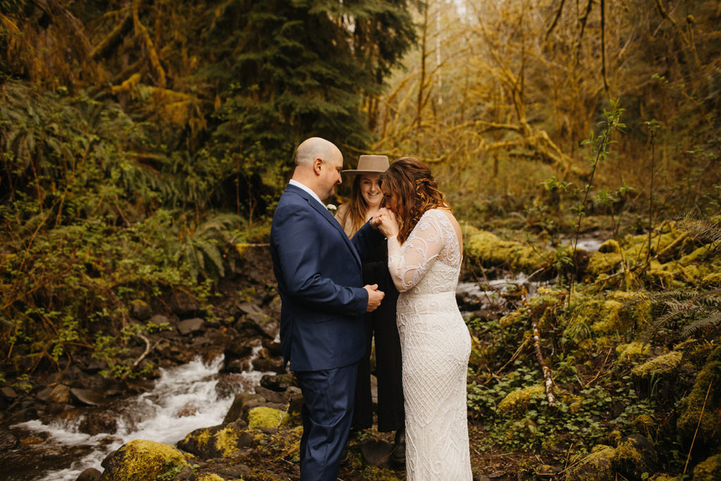 rain forest elopement bride kisses grooms hand while officiant smiles in background